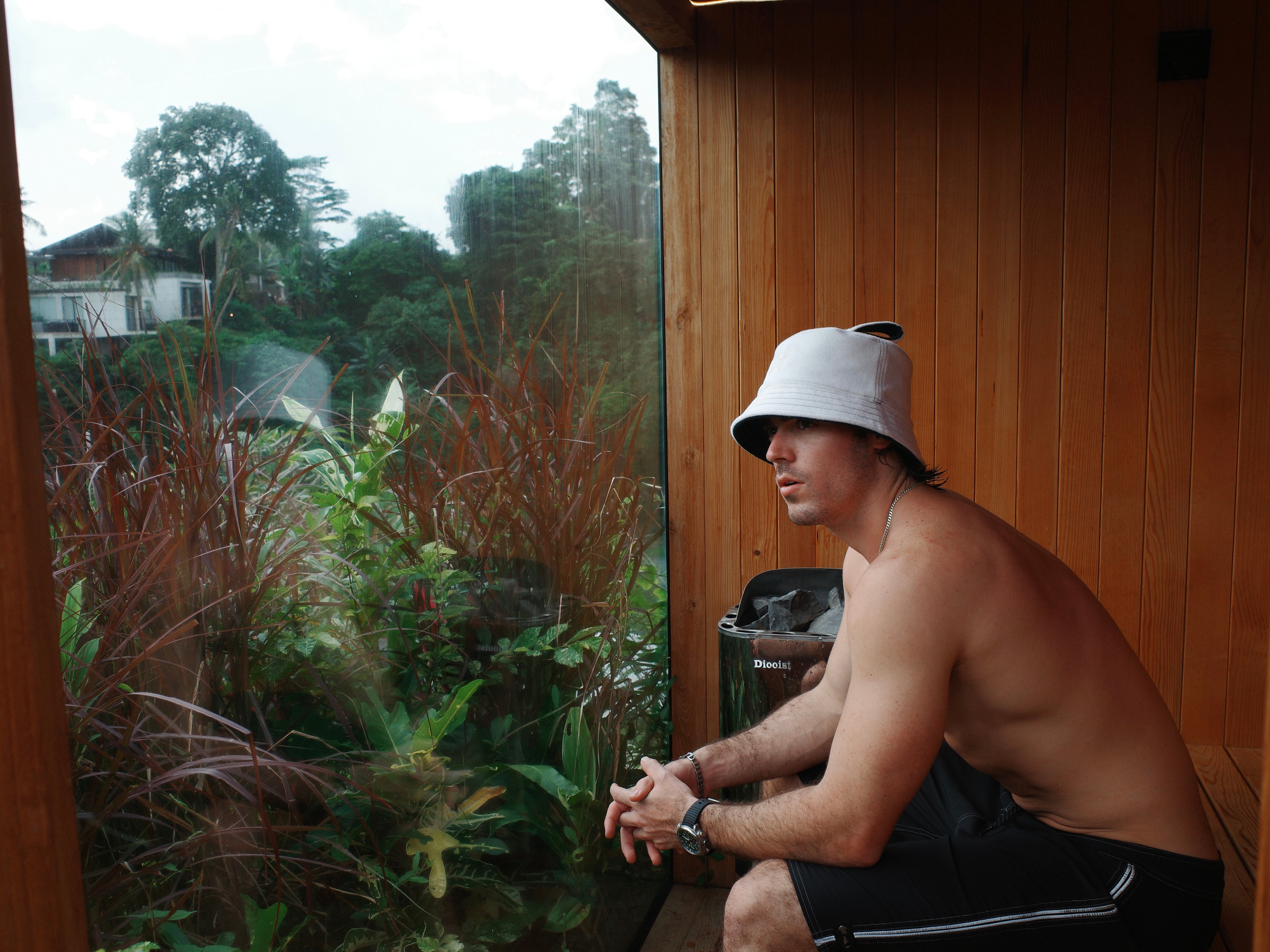 Man sitting in wooden sauna wearing a Halo sauna hat looking out glass window