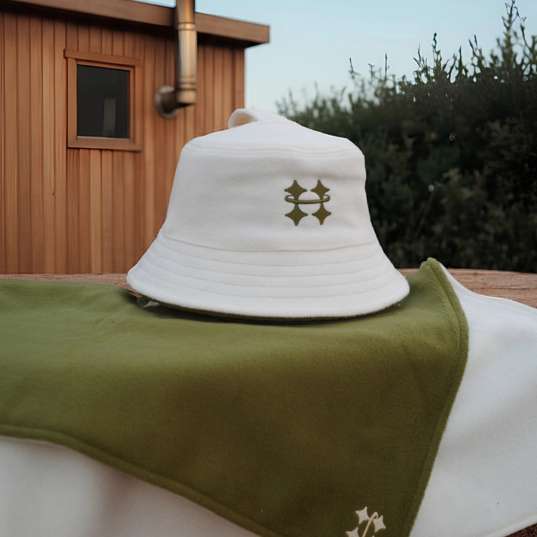A white Halo sauna hat with a olive green logo sits on top of an olive green sauna mat outdoors, with a wooden cabin and greenery in the background.