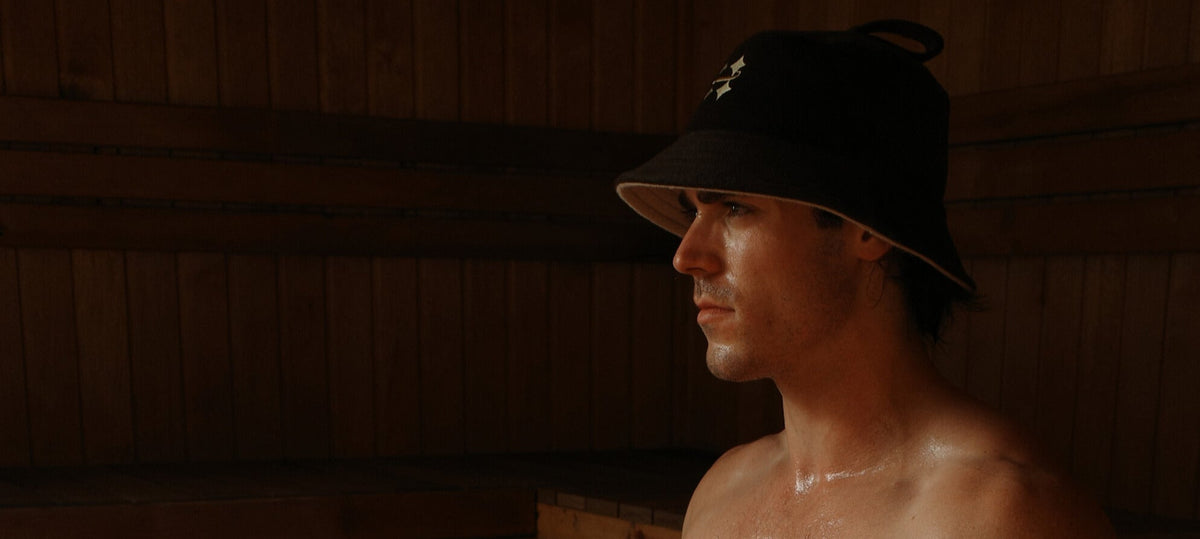 A man wearing a brown Halo sauna hat with a small embroidered logo, sitting in a dimly lit wooden sauna.